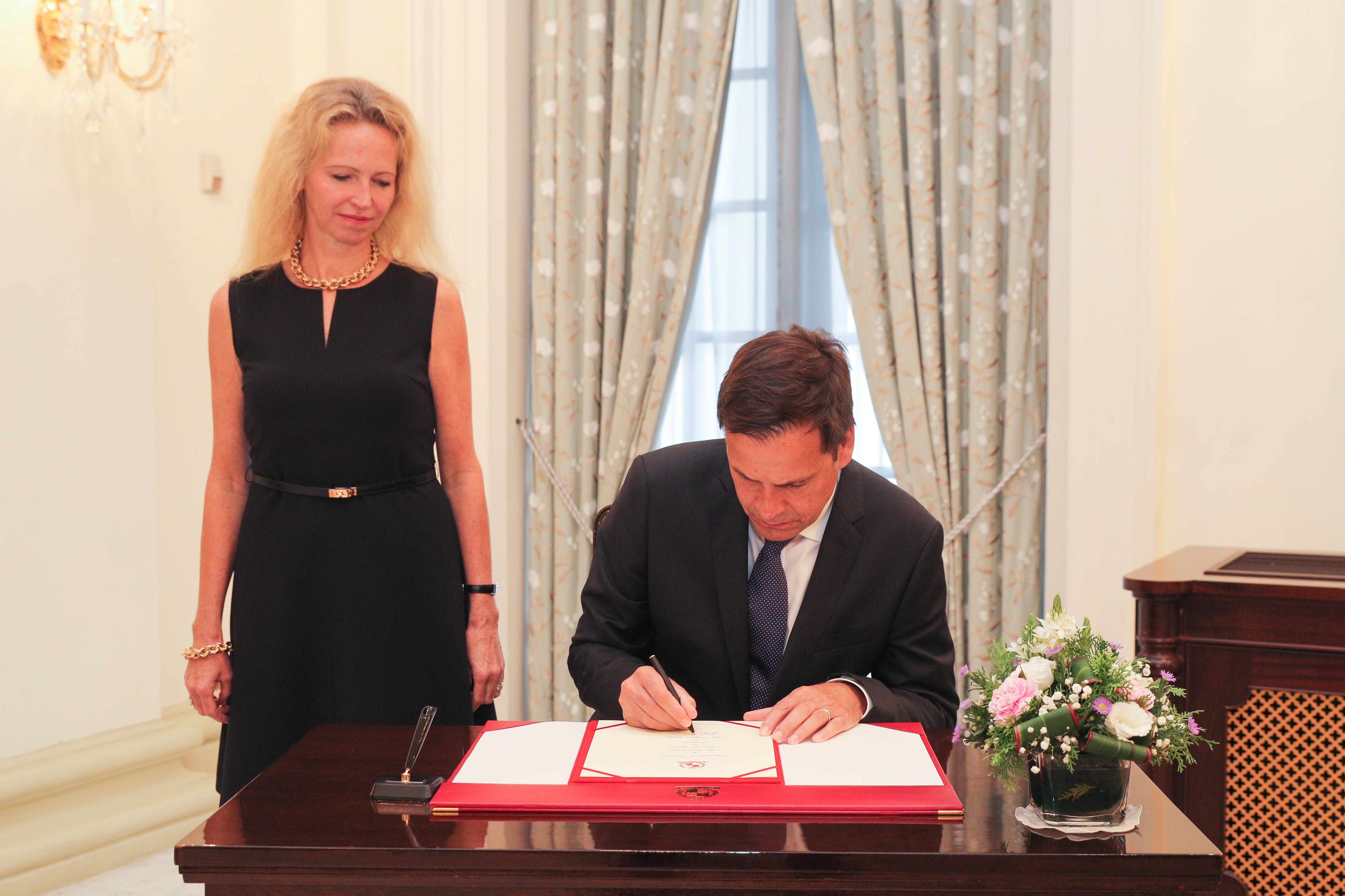 Man signs document on red blotter. Woman stands nearby. Floral arrangement on table.